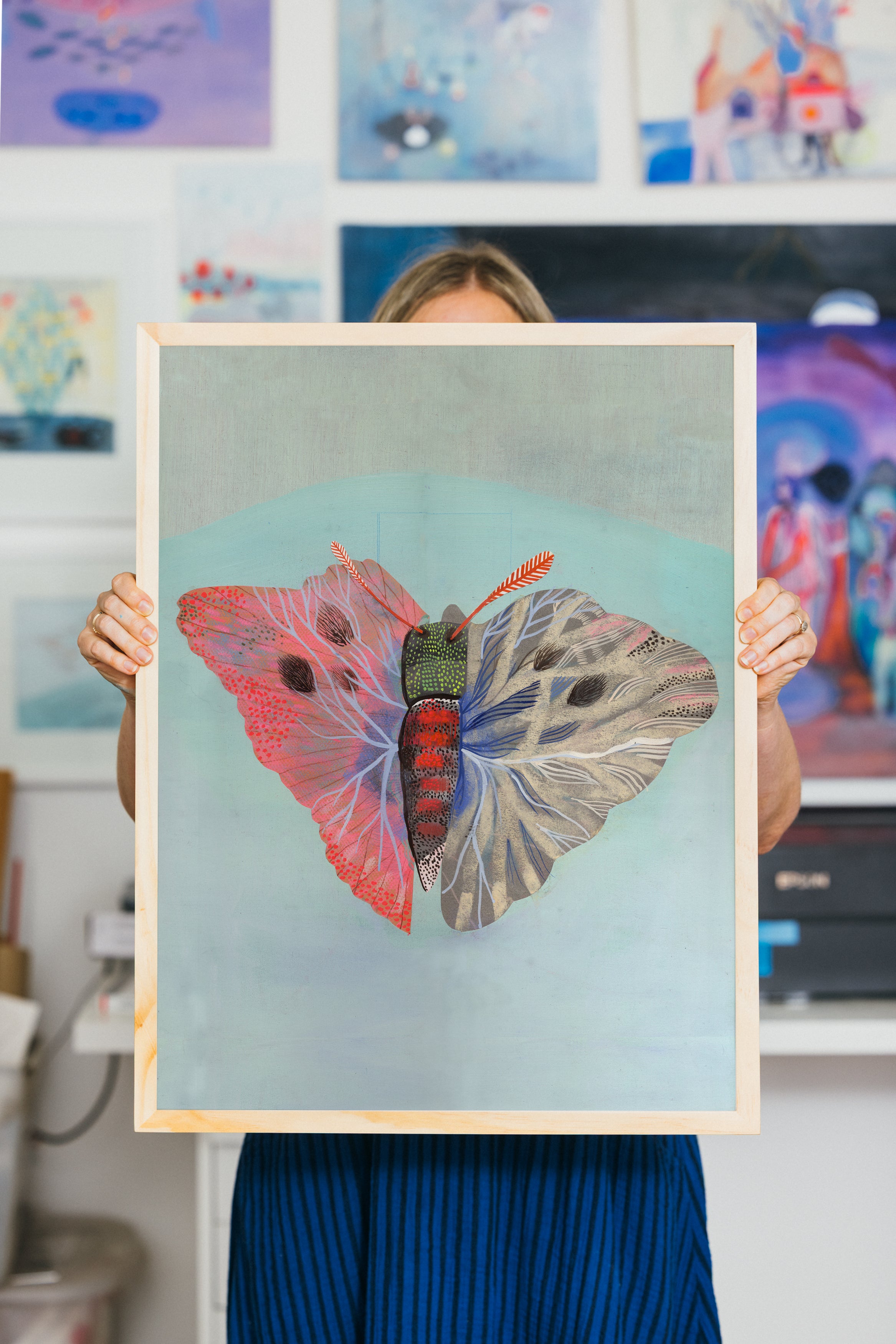 Large butterfly art print held by artist in a studio setting, showing detailed pastel and painted textures in vivid and neutral tones.