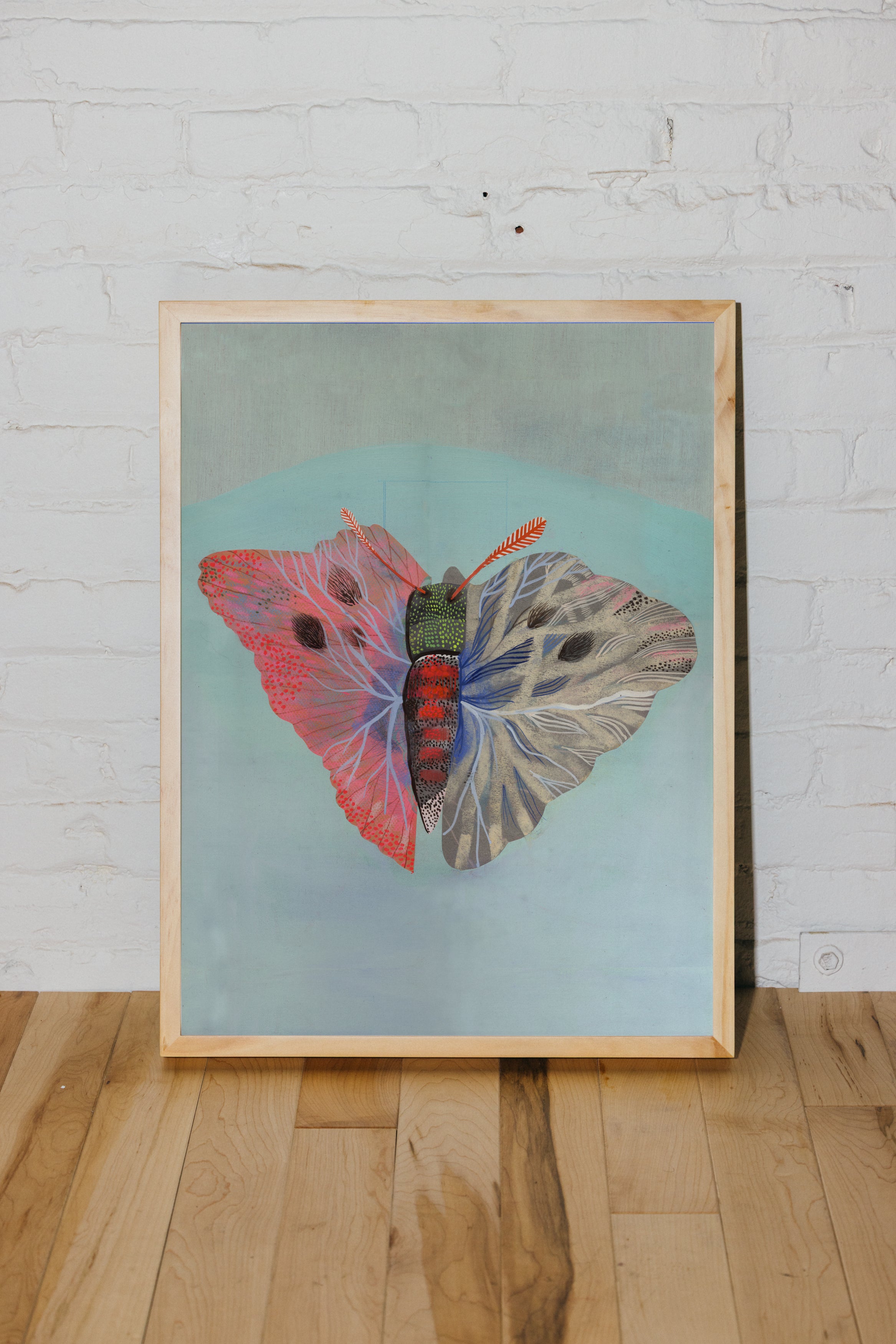 Archival 17x22 giclée art print of a butterfly painting by Betsy Walton, displayed in a natural wood frame against a white brick wall.